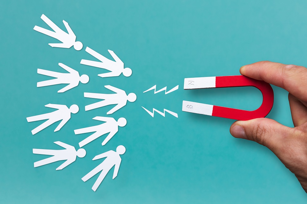 Close-up Of A Human Hand Attracting White Human Figures With Horseshoe Magnet On Blue Background. Representing Lead Generation